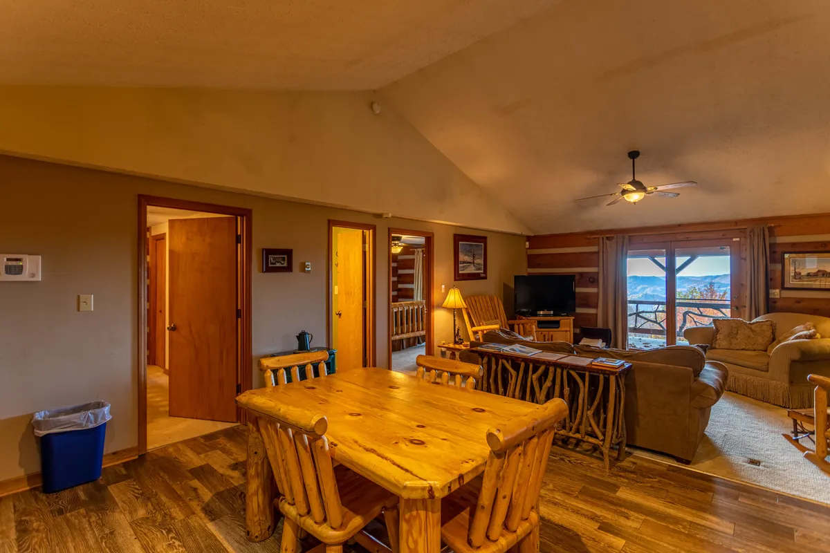 This interior image showcases a wooden dining table adjacent to a cozy sofa and a TV, with a large window that lets in natural light and offers scenic views. The setting blends dining and relaxation, supporting both meal times and extended gatherings.