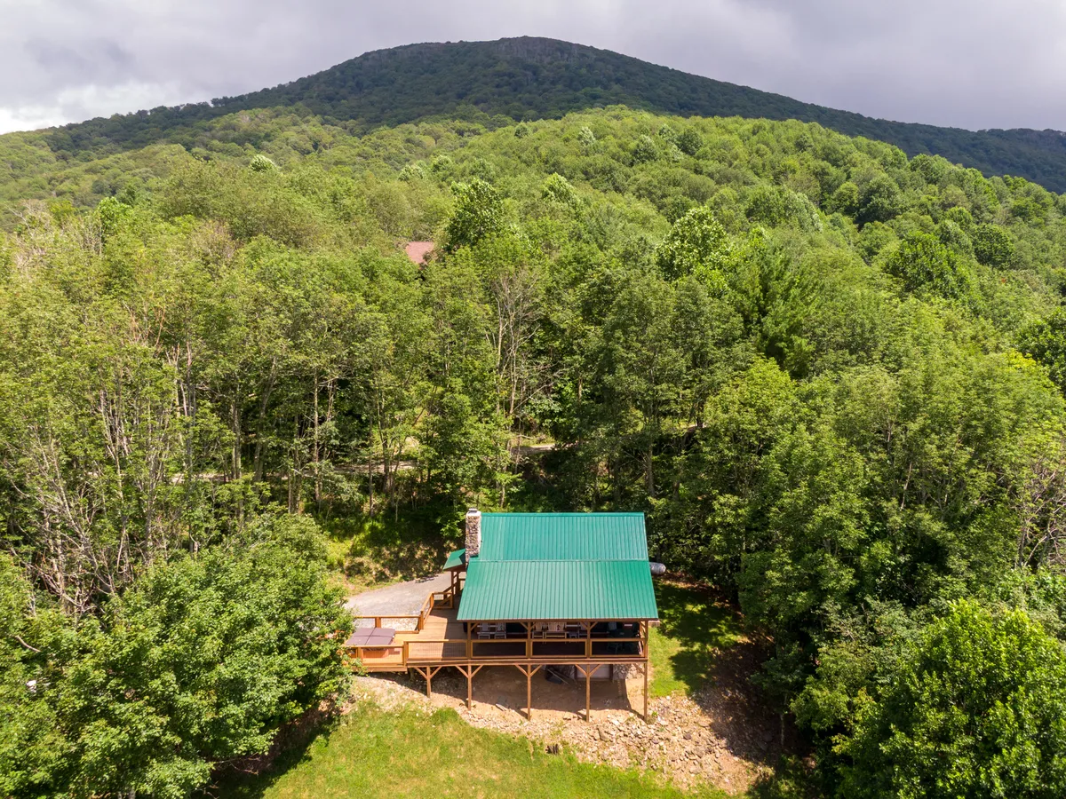 This exterior shot shows the house nestled among trees with a dramatic mountain backdrop, highlighting the porch where guests can relax and take in the peaceful scenery. The image evokes a sense of seclusion, privacy, and connection with nature, perfectly matching the retreat's romantic appeal.
