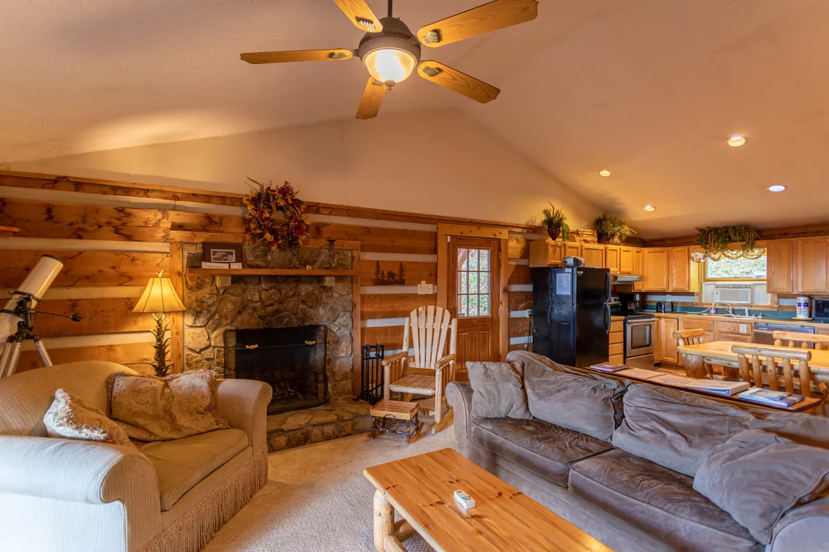 A rustic-chic living room featuring a stone fireplace, comfortable wooden furniture, and large windows that flood the space with natural light. The seating arrangement is perfect for movie nights or family gatherings, and the atmosphere is both welcoming and tranquil, with mountain views visible in the background.