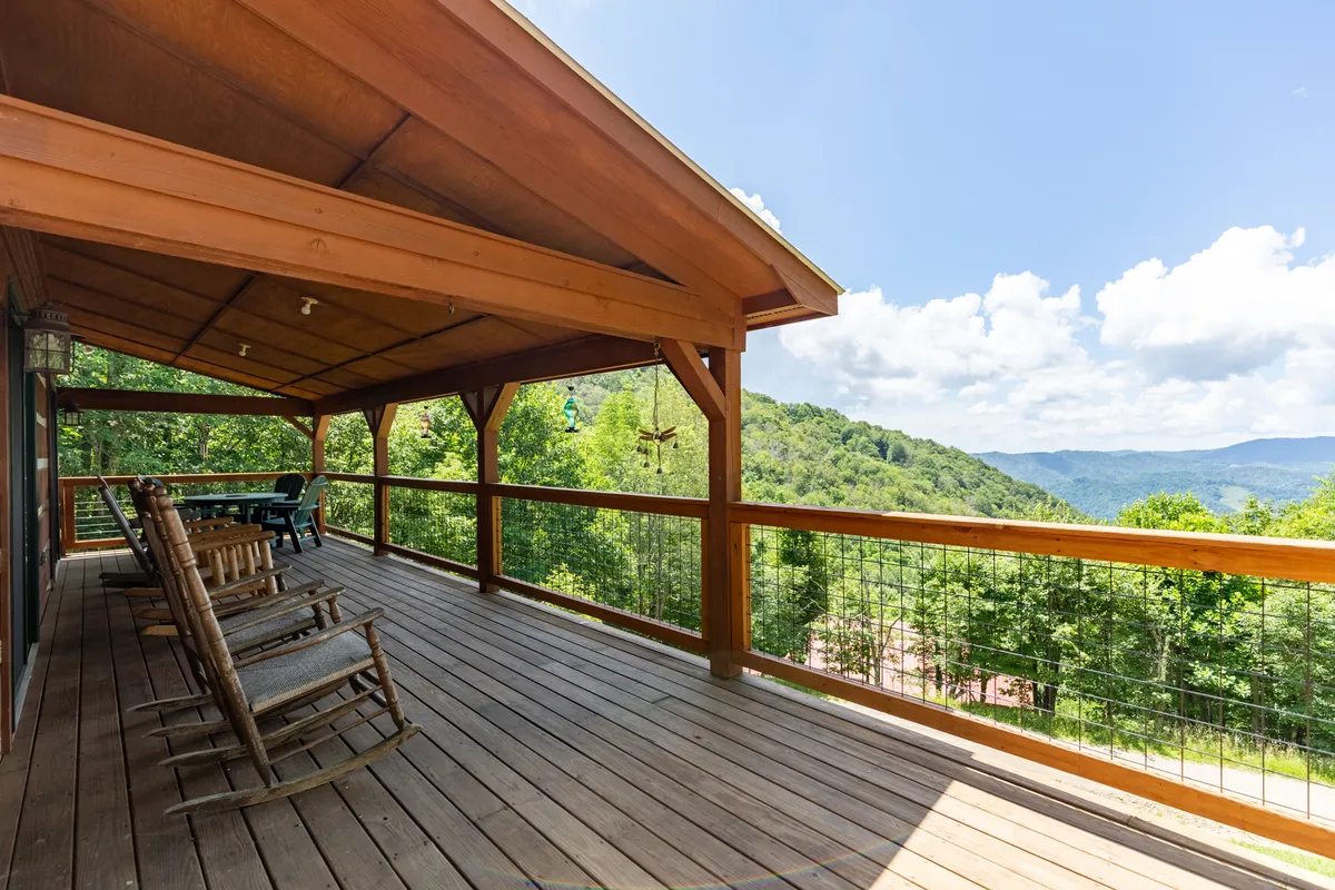 This image showcases a classic covered wooden porch lined with rocking chairs, offering unobstructed panoramic views of distant mountains framed by a sturdy wooden railing. The tranquil scene is set for relaxation, with the porch extending along the home's length and nature unfolding just beyond its edge.