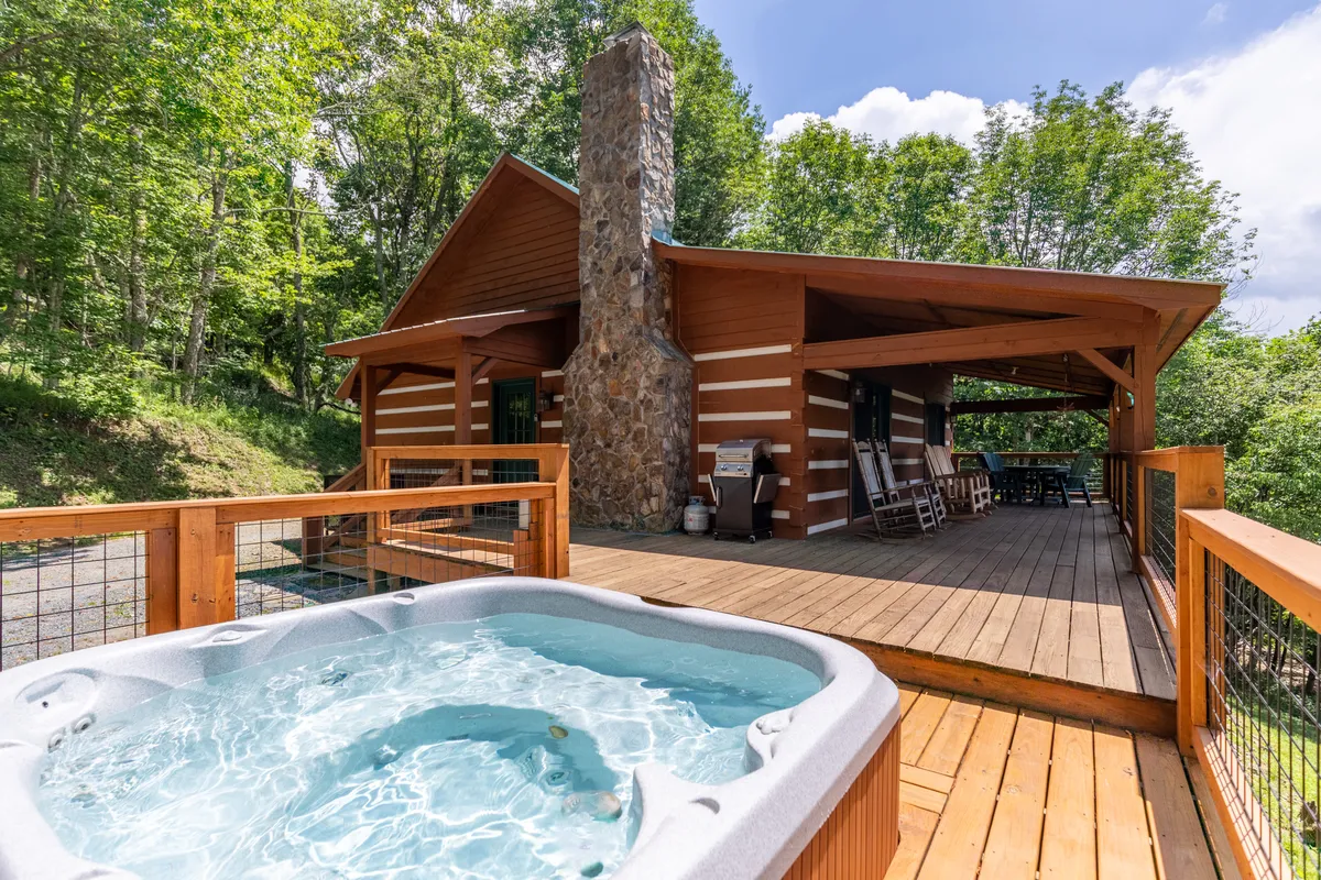 This image features the log cabin’s stone chimney and wooden deck, with the hot tub as a focal point. The setting is tranquil, surrounded by nature—perfect for both relaxing and socializing. The deck’s rocking chairs and panoramic windows hint at inviting workspaces and spots for coffee breaks while soaking in the peaceful environment.