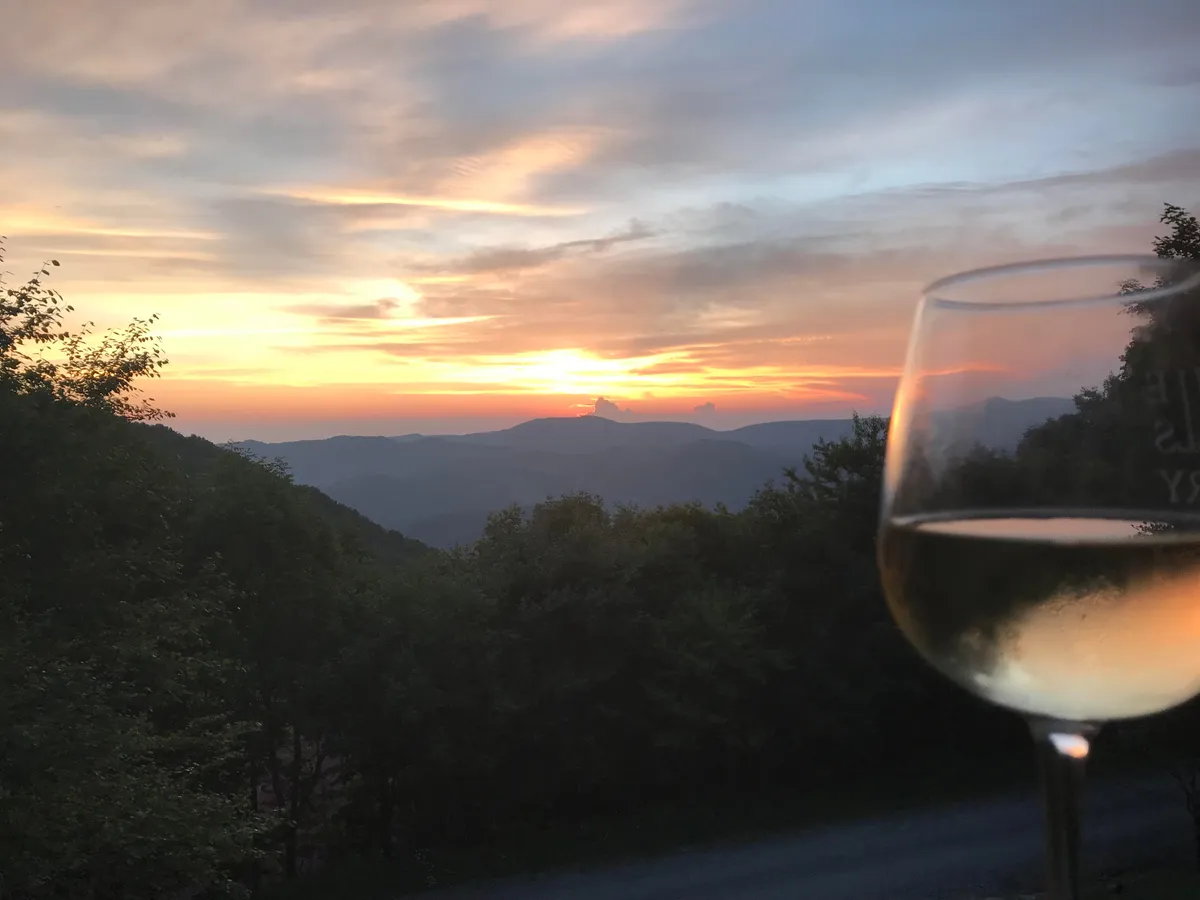 A breathtaking view of a fiery sunset over the mountains, with a glass of wine held in the foreground, suggesting a moment of quiet appreciation and celebration. The scene is serene, intimate, and bathed in warm evening light.