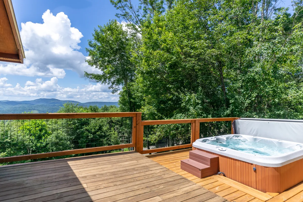 A wide view of the cabin’s elevated wooden deck, featuring a hot tub overlooking sweeping mountain vistas. The scene includes the deck railings, comfortable seating, and an open sky—suggesting the transition from indoor to outdoor adventure, with the mountains as the backdrop.