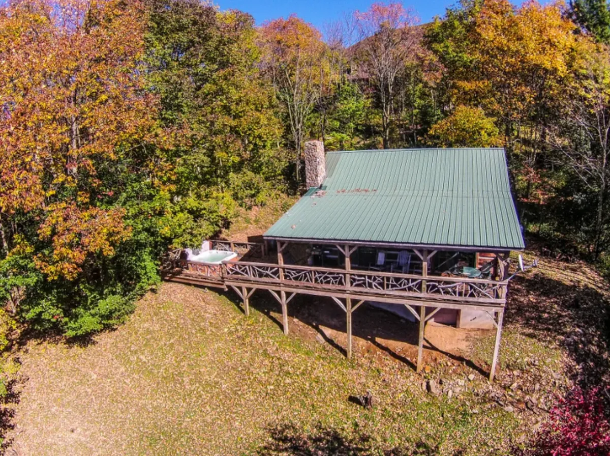 A wide shot of the cabin’s wooden deck, green metal roof, surrounding trees, and a prominent hot tub. The image captures the sense of escape and privacy, with the deck set up for relaxation, meals, and stargazing—all with mountain air and a connection to the outdoors.