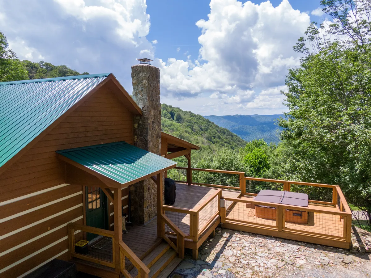 The image showcases a classic log cabin with a striking stone chimney, nestled atop a ridge. The spacious deck features a bubbling hot tub overlooking breathtaking, expansive mountain views. The setting sun bathes the scene in golden light, inviting guests to unwind and savor the tranquility.
