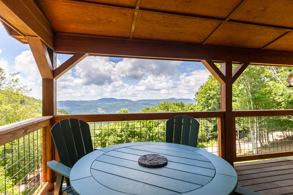 A spacious wooden deck furnished with a circular table and Adirondack chairs overlooks sweeping mountain views. The scene is serene, with the promise of gathering for meals, morning coffee, or evening stargazing while surrounded by nature.