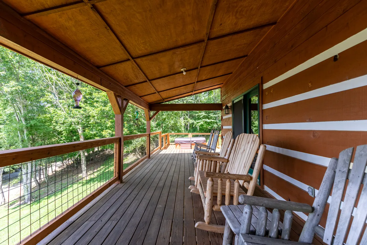 A comfortable stretch of covered porch with wooden railings features inviting rocking chairs and a hanging lantern, overlooking a lush green landscape. The setup suggests a perfect spot for sipping drinks, relaxing, or enjoying conversation as the sun sets.