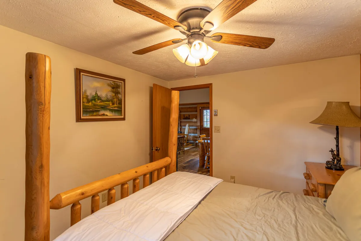 A warmly lit bedroom centered on a log bed frame, ceiling fan, and wooden lamp. The space feels open and welcoming, with an open door leading to another room, suggesting connectivity within the suite.