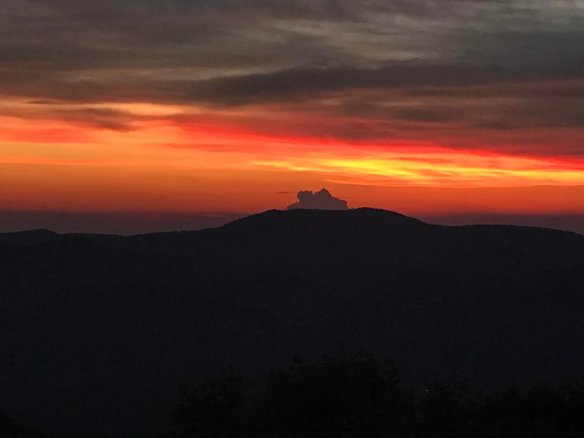 A dramatic sunset washes the sky in vibrant hues above silhouetted mountains, with a few wispy clouds adding depth. The wide, open view creates a sense of awe and tranquility, perfectly encapsulating the magic of Appalachian evenings and the allure of nature's nightly display.