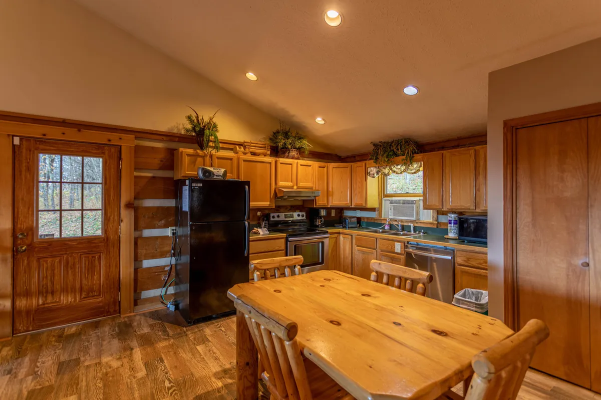 This image features a wooden dining table set in an inviting kitchen space, with a black refrigerator and kitchen cabinets in the background. Soft natural light streams in from a window, adding warmth and a sense of openness. The setup highlights a space designed for gathering and meal preparation.
