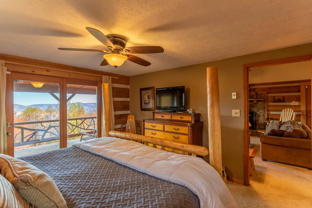 A cozy bedroom featuring a comfortable bed positioned near windows that offer a view of the mountains. The room includes a wooden dresser, draped curtains, and a wall-mounted television, all arranged to maximize both comfort and scenic enjoyment.