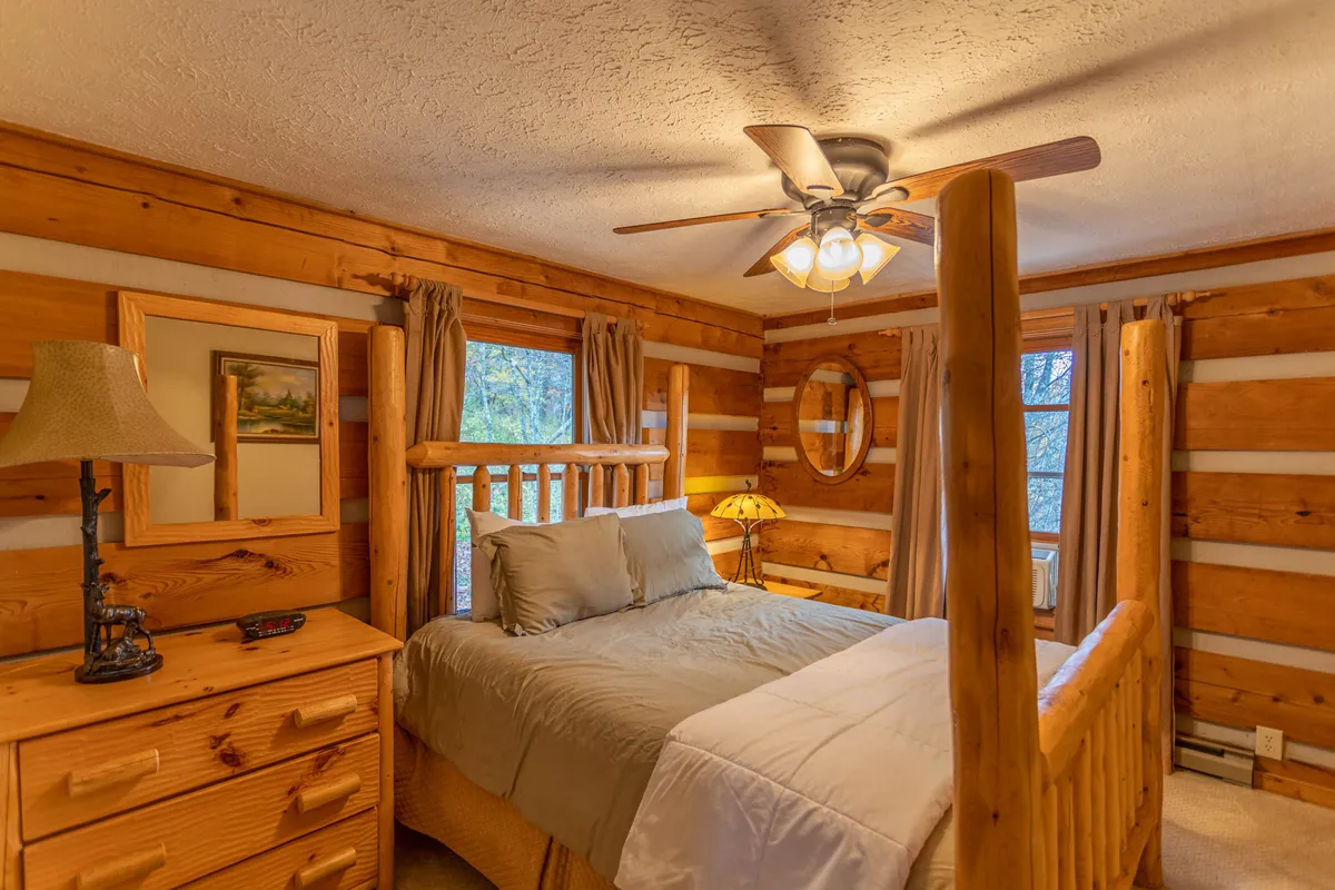 An inviting bedroom with a wooden four-poster bed, matching dresser, soft lighting from a bedside lamp, and framed artwork, all evoking a rustic, cabin-inspired ambiance.