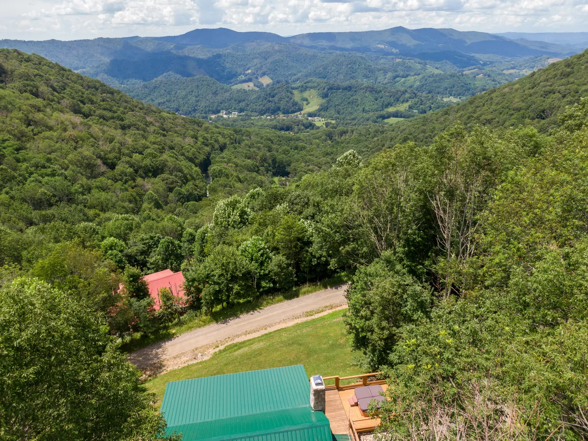 A breathtaking landscape shot of the surrounding High Country, featuring rolling mountains, a wooded valley, and a winding road leading toward adventure. The image also includes a red-roofed building nestled in the trees, emphasizing the remote, nature-rich environment.