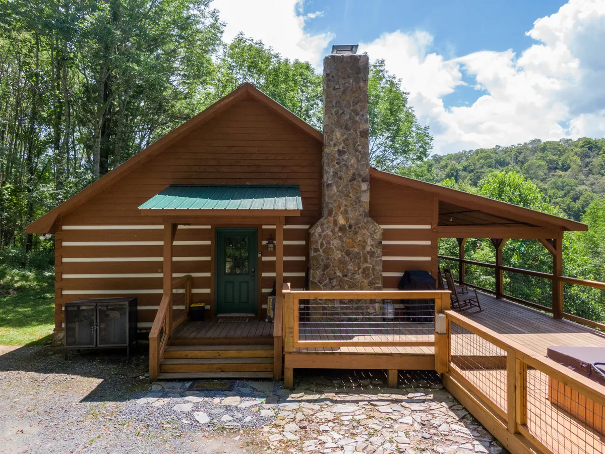 The cabin’s entrance and deck area greet visitors with a rustic log structure, stone chimney, and inviting outdoor spaces. The setting is bathed in warm light, suggesting the perfect spot to transition from work to relaxation as the sun sets.