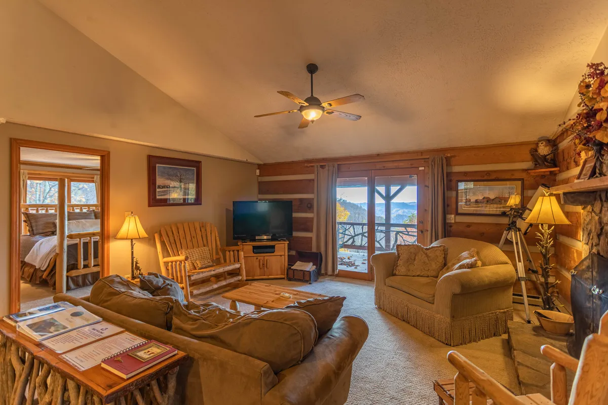 This image displays the inviting living room with warm wooden furniture, a prominent stone fireplace, a television, and large windows that fill the space with natural light. The arrangement of plush seating creates a welcoming and comfortable atmosphere, perfect for relaxing, socializing, or enjoying an evening by the fire. The large glass windows showcase the stunning outdoor scenery, reinforcing the connection to the mountains.